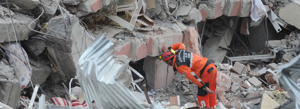 Slika člana reševalne ekipe v oranžnem kombinezonu, ki preiskuje betonske ruševine.