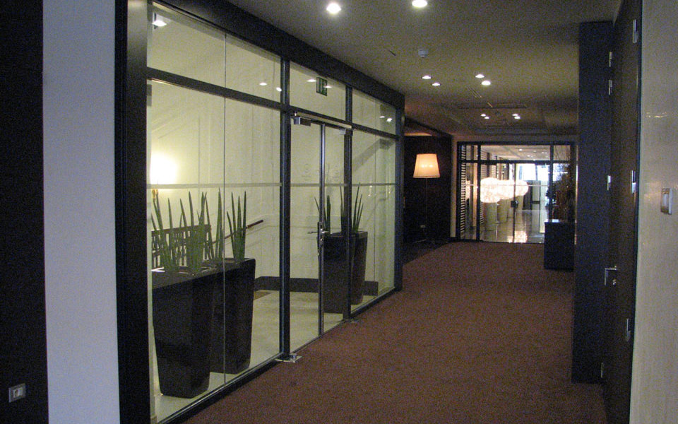 Na sliki je hodnik, kjer je uporabljena požarna zasteklitev brez profilov Promat®-SYSTEMGLAS 30 v notranjosti hotela Kempinski Palace v Portorožu, na levi strani zasteklitve so zelene rastline v velikih loncih. 