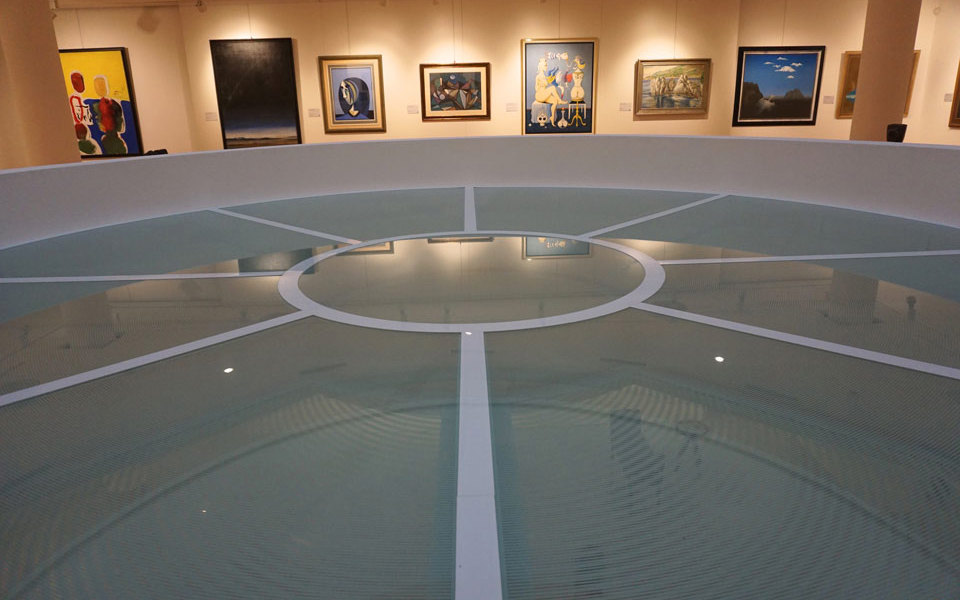 Na sredini slike je okrogla horizontalno zastekljena konstrukcija s požarnimi stekli PROMAGLAS® v prosotru galerije Nedbalka v Bratislavi, v ozadju je razstava umetniških slik v galeriji. 