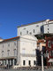 Pogled iz Tartinijevega trga, levo je spomenik Tartiniju, na sredini je bela zunanjost Mestne galerije Pirana, desno so rdeča, bela in rumena hiša na trgu. 