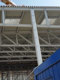 Jeklene konstrukcije na začetku gradnje dvorane Gimnastičnega centra Ljubljana. Konstrukcije so bele barve, nad njimi vidimo del žerjava, pod njimi pa del modrih kontejnerjev za delavce.   