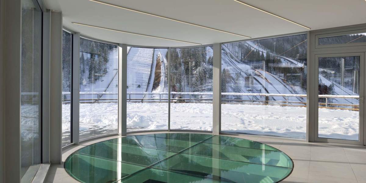 Steklena notranjost zgornjega nadstropja objekta Nordijskega centra Planica z okroglim horizontalnim pohodnim steklenim oknom v tleh. V ozadju se skozi stekla vidijo smučarske skakalnice v snegu. 