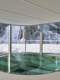 Steklena notranjost zgornjega nadstropja objekta Nordijskega centra Planica z okroglim horizontalnim pohodnim steklenim oknom v tleh. V ozadju se skozi stekla vidijo smučarske skakalnice v snegu. 
