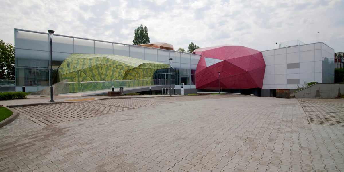 Zunanjost muzeja Muzeiko, ki je ogromen podolgovat steklen objekt, z dvema izbočenima modernima arhitekturama oziroma izboklinama. Leva je zelena, desna je rdeča. Pred muzejem so rdeče tlakovana tla. 