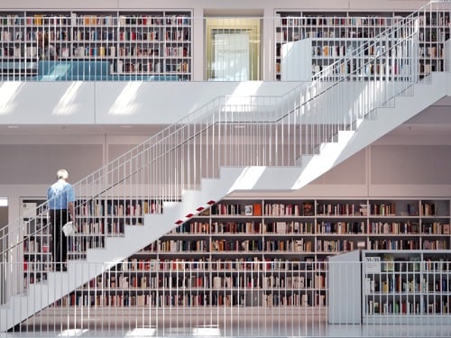 Notranjost knjižnice z belo konstrukcijo in belimi stopnicami, ki gredo diagonalno v zgornje nadstropje, na njih stoji oseba. V ozadju so knjige. 