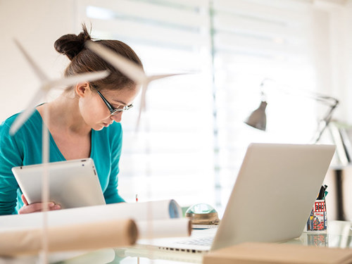 Žena sa slike u plavoj majici sjedi za stolom i gleda papire, ispred nje je bijeli laptop i bijeli smotuljci papira. U pozadini je stolna lampa.