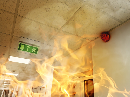 Slika zgornjega dela hodnika in ognjeni zublji, na stropu je zelen napis za izhod v sili, na desni strani slike pa naprava za zaznavo dima. 