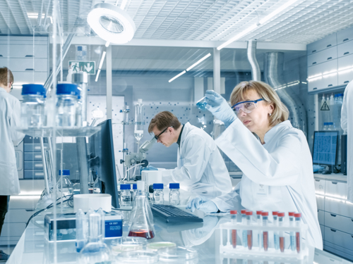 Četvorica znastvenika u bijelim haljinama u laboratoriju. U glavnom kadru je znanstvenica koja promatra epruvetu i znanstvenik koja gleda u mikroskop.