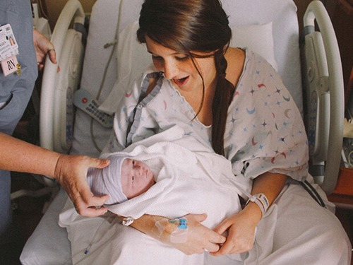 Slika prikazuje mati, ki sedi v postelji v porodnišnici in se navdušeno smeje ter drži v rokah svojega novorojenega otroka. 