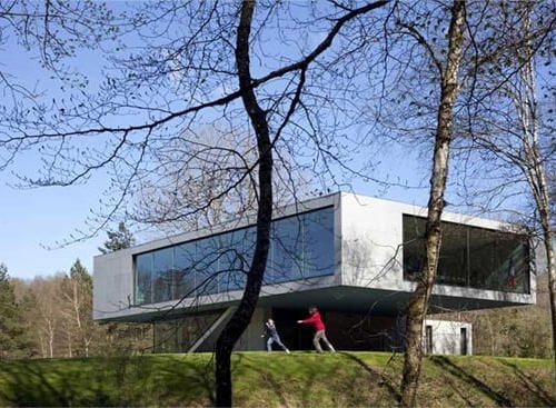 Na slici je iza drveća moderna kuća u prirodi sa ostakljenim zidovima. Ispred kuće dvije osobe vježbaju.