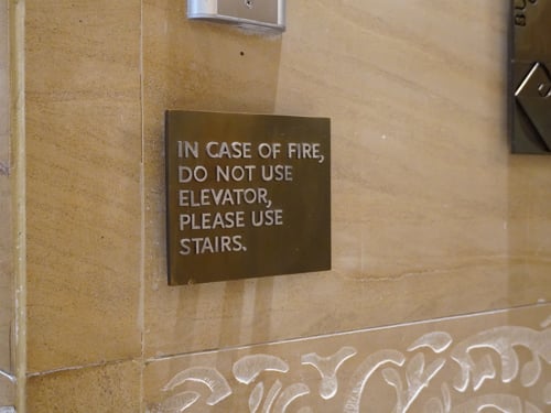 Na napisu na rjavi plošči na steni s svetlo rjavimi ploščicami je zapisano: In case of fire, do not use elevator, please use stairs.  