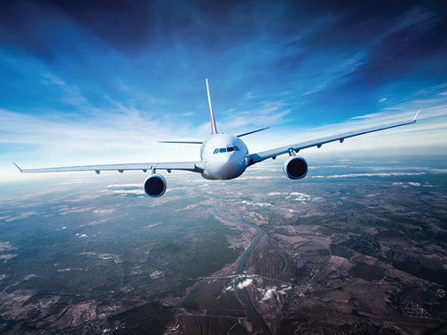 Belo letalo slikano iz profila, ki leti visoko v zraku. Nebo okoli letala je svetlo modro, pod letalom pa se vidi rjavo zelena površina zemlje. 
