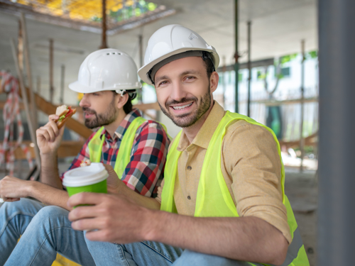 Dobro raspoloženi radnici u belim šlemovima i fluorescentnim prslucima prave pauzu za užinu u toku rada na građevini.