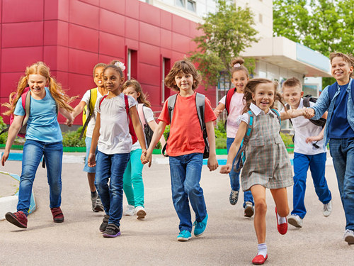 Skupina veselih otrok hodi po ulici, v ozadju se vidi zgradbo šole v rdeči in beli barvi. 