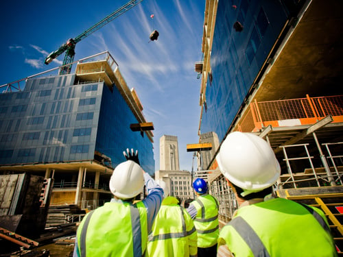 Štirje inženirji v rumenih odsevnih jaknah s čeladami na glavi, opazujejo gradnjo dveh modro zastekljenih zgradb. Nad levo stavbo je žerjav v gibanju, ki prenaša tovor. 