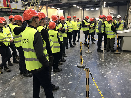 Velika skupina inženirjev v rumenih odbojnih brezrokavnikih s črnimi rokavi in rdečimi čeladami na glavi, stoji in posluša razlago v industrijskem prostoru. 