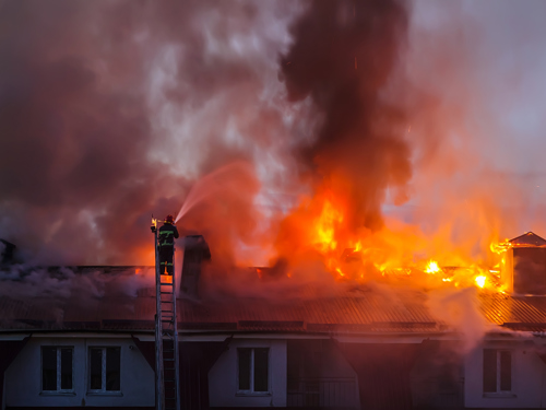 Vatrogasac na merdevinama gasi požar koji bukti na krovu stambenog objekta vodom iz vatrogasnog creva dok se tamni dim diže.