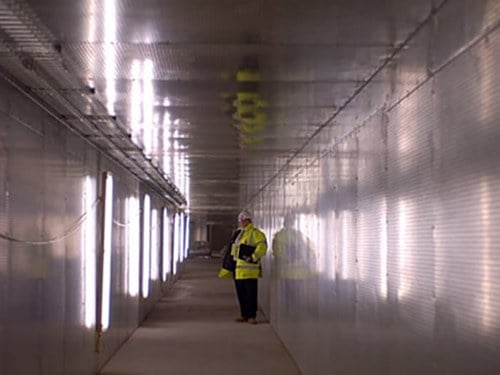 Inspektor u žutoj jakni i sa šlemom na glavi vrši inspekciju protivpožarne zaštite dugačkog hodnika građevinskog objekta.