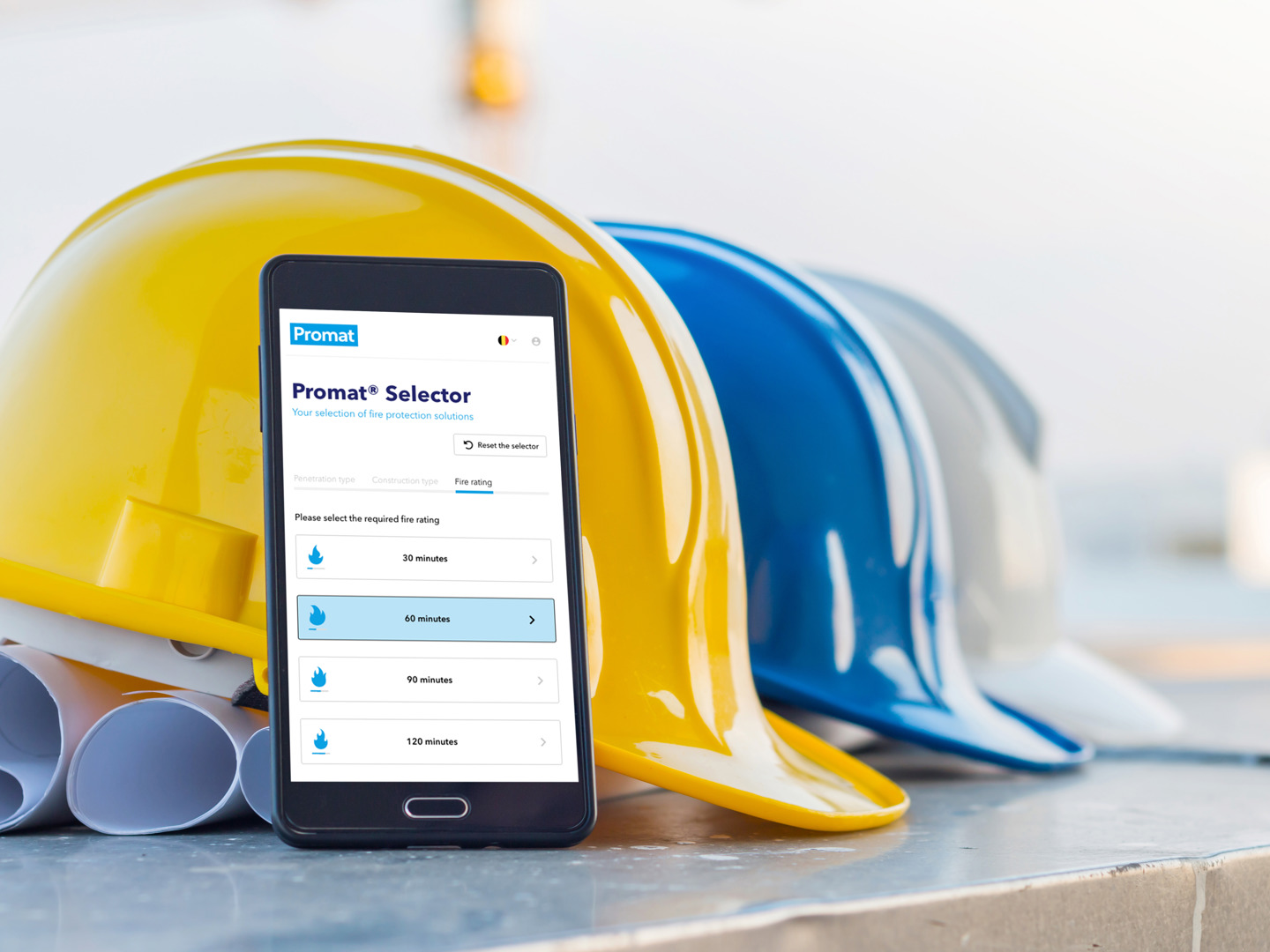Trei căști de protecție de diferite culori pe o masă cu niște planuri și un telefon cu aplicația Promat Selector deschisă.