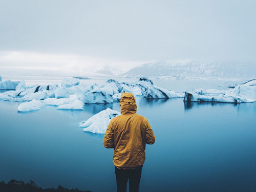 Persoană îmbrăcată în galben stând lângă apă într-un peisaj muntos cu gheță
